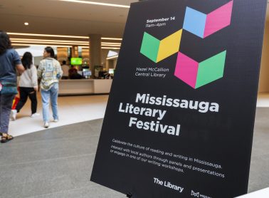A large black promotional sign for the Mississauga Literary Festival stands inside Hazel McCallion Central Library. The sign features colourful geometric shapes and festival information, while several visitors walk through the bright, modern library lobby in the background.