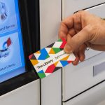 A hand holding up a Mississauga Library card with a digital library self-checkout machine in the background