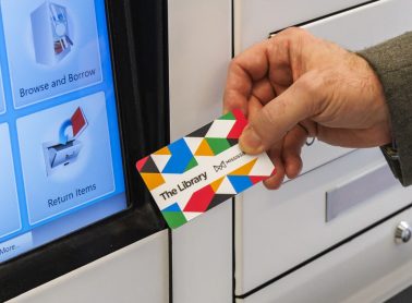 A hand holds a Mississauga Library card beside a self-service library kiosk screen. The screen displays options for borrowing and returning library materials.