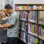 A library worker helps a customer using a tablet, they are standing in front of bookshelves
