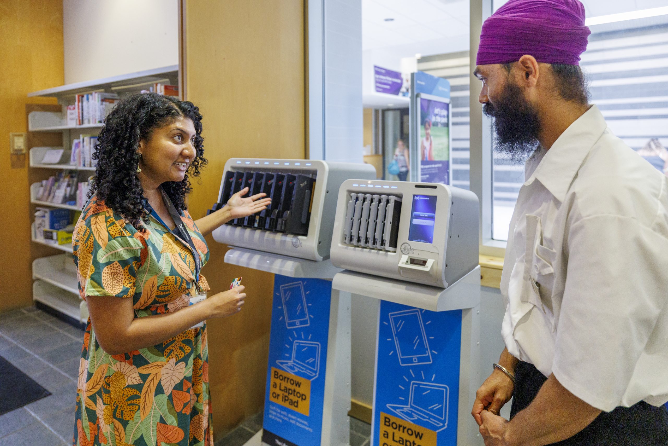 A library worker is showing a customer how to sign out technology while they stand in front of a shelf with laptops and tablets. There are standing in a bright, open space with bookshelves in the background.