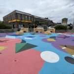 Colourful ground murals with wood seating on the corner of a retail plaza and busy street.