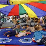 Caregivers and infants participate in an early literacy program at a library, sitting on a colourful alphabet rug while holding up a large multicoloured parachute overhead. Babies play with toys and look up at the parachute in an open indoor community space.