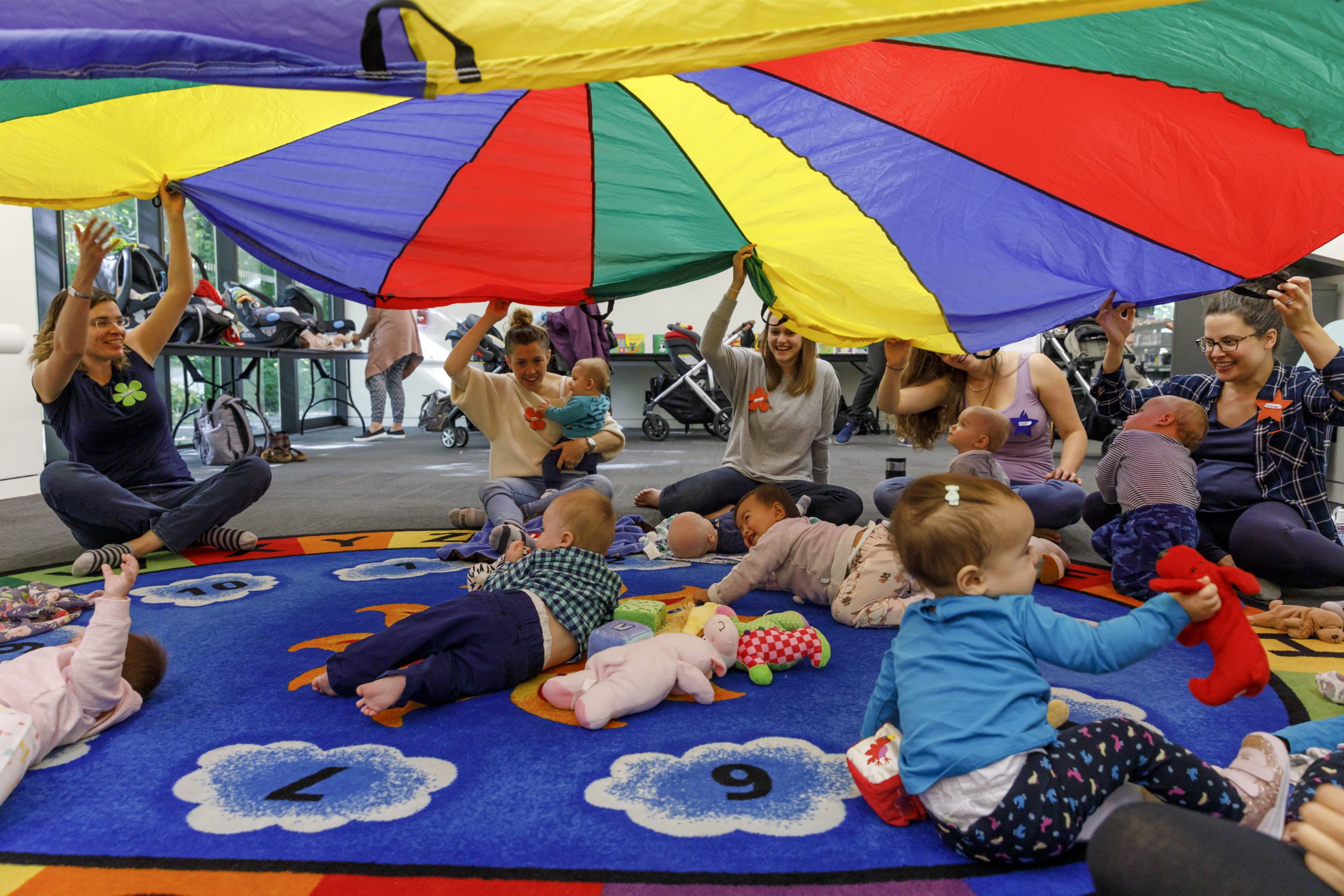 Caregivers and infants participate in an early literacy program at a library, sitting on a colourful alphabet rug while holding up a large multicoloured parachute overhead. Babies play with toys and look up at the parachute in an open indoor community space.
