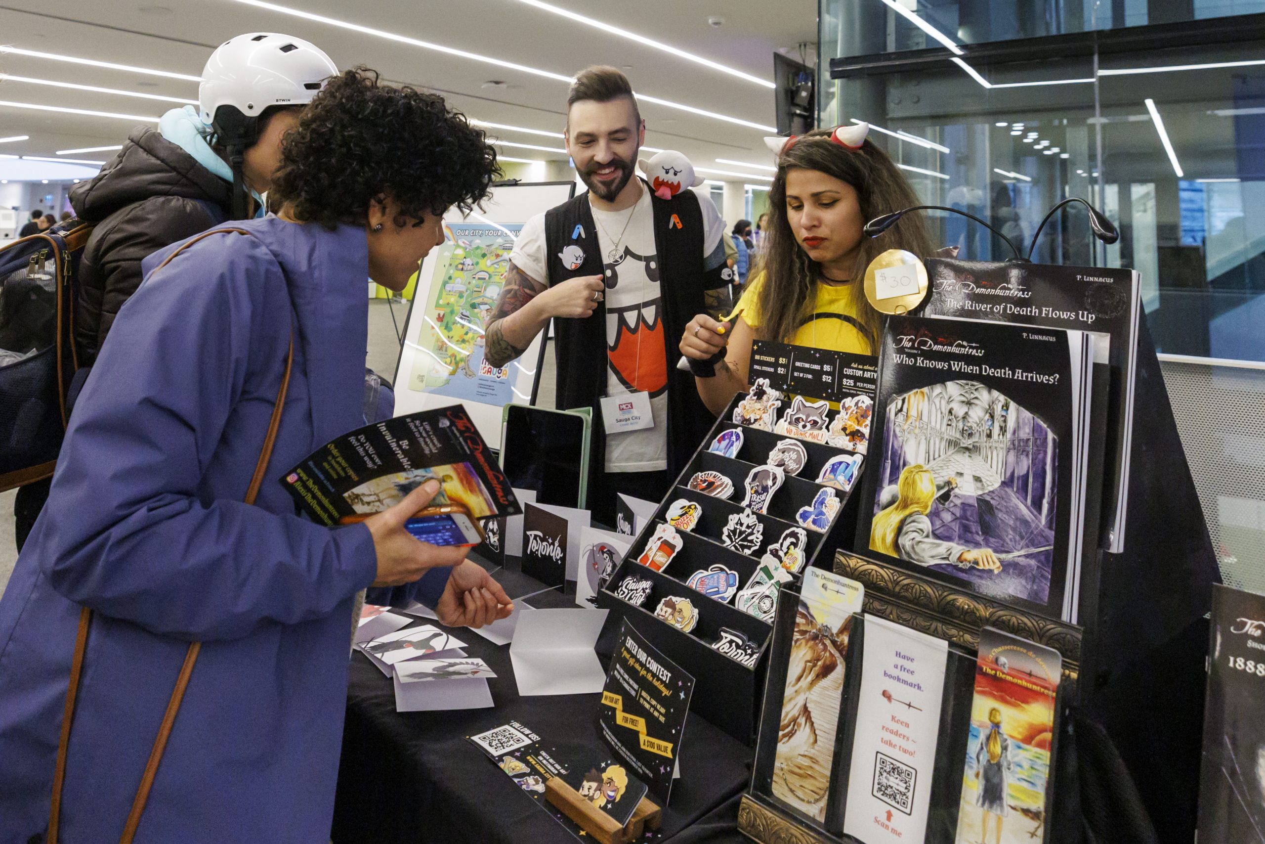 Visitors browse a vendor table displaying books, artwork, stickers and printed materials at a community event in a modern library space. Several people are speaking with exhibitors and exploring locally created publications and merchandise.