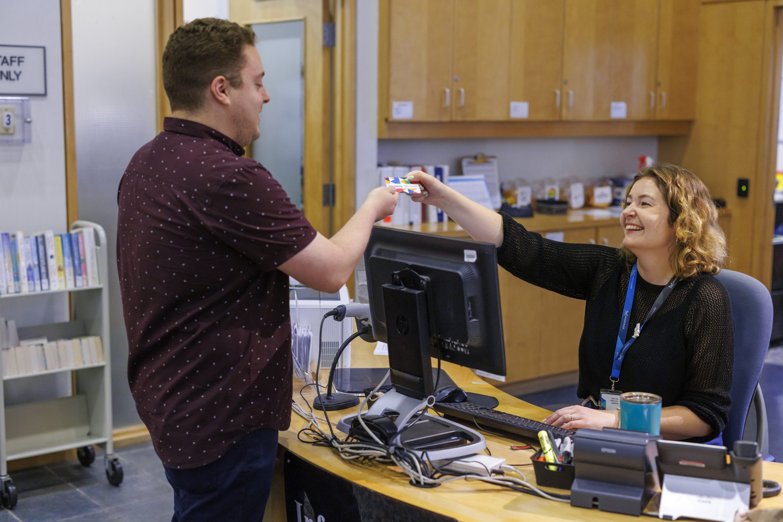 A library staff member hands a library card to a visitor at a service desk. Computers, library materials and shelving are visible in the background of the library branch.