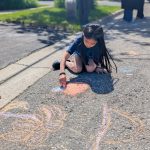 A girl draws with sidewalk chalk on the pavement on a road that has been closed to vehicle traffic.