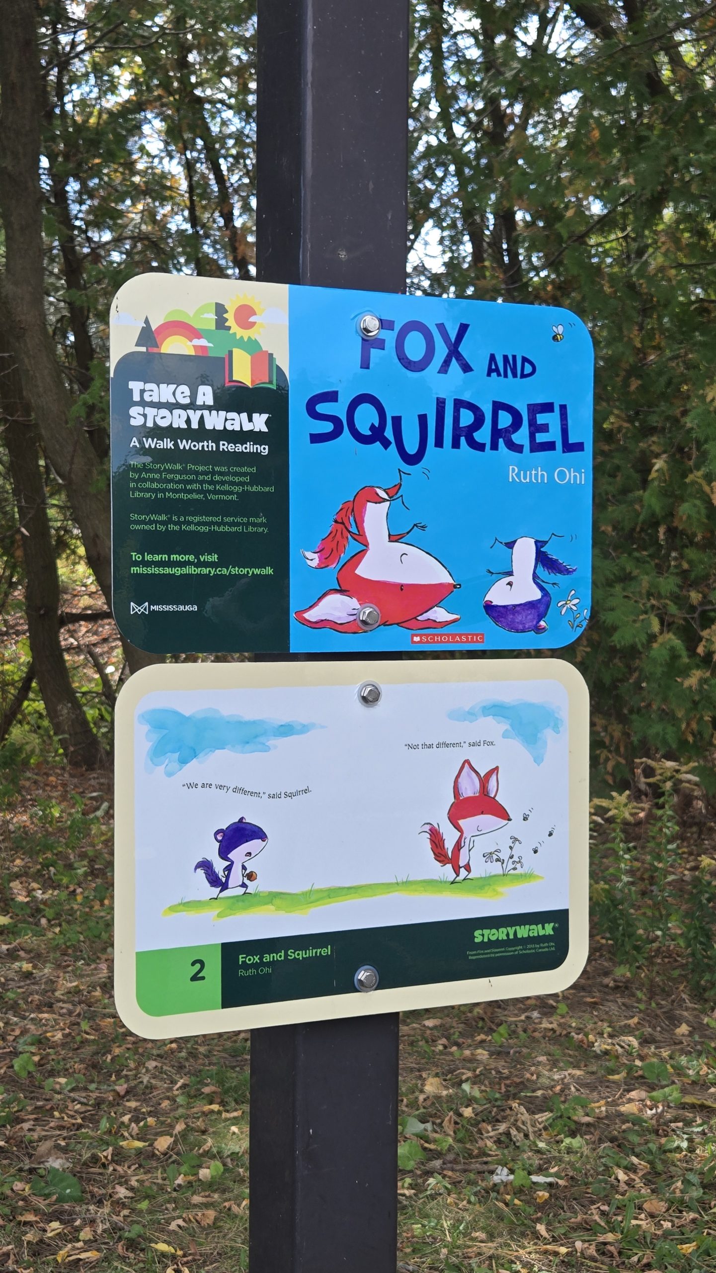 StoryWalk sign mounted on a post in a wooded park. The top panel displays the cover of the children's book Fox and Squirrel by Ruth Ohi. The lower panel shows the first page of the story, featuring illustrated fox and squirrel characters facing each other on a grassy area, surrounded by trees and foliage in the background.