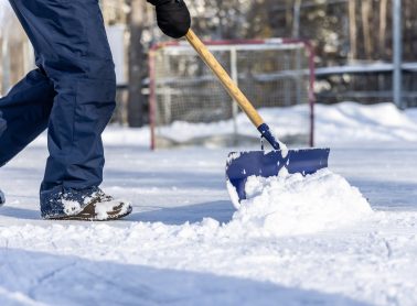 A man is shovelling snow on an outdoor ice rink before a hockey game. He is working very hard to clear the ice rink. It is a beautiful sunny winter day.