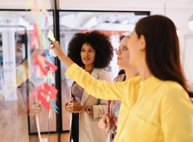Close-up of women putting sticky notes on a glass wall.