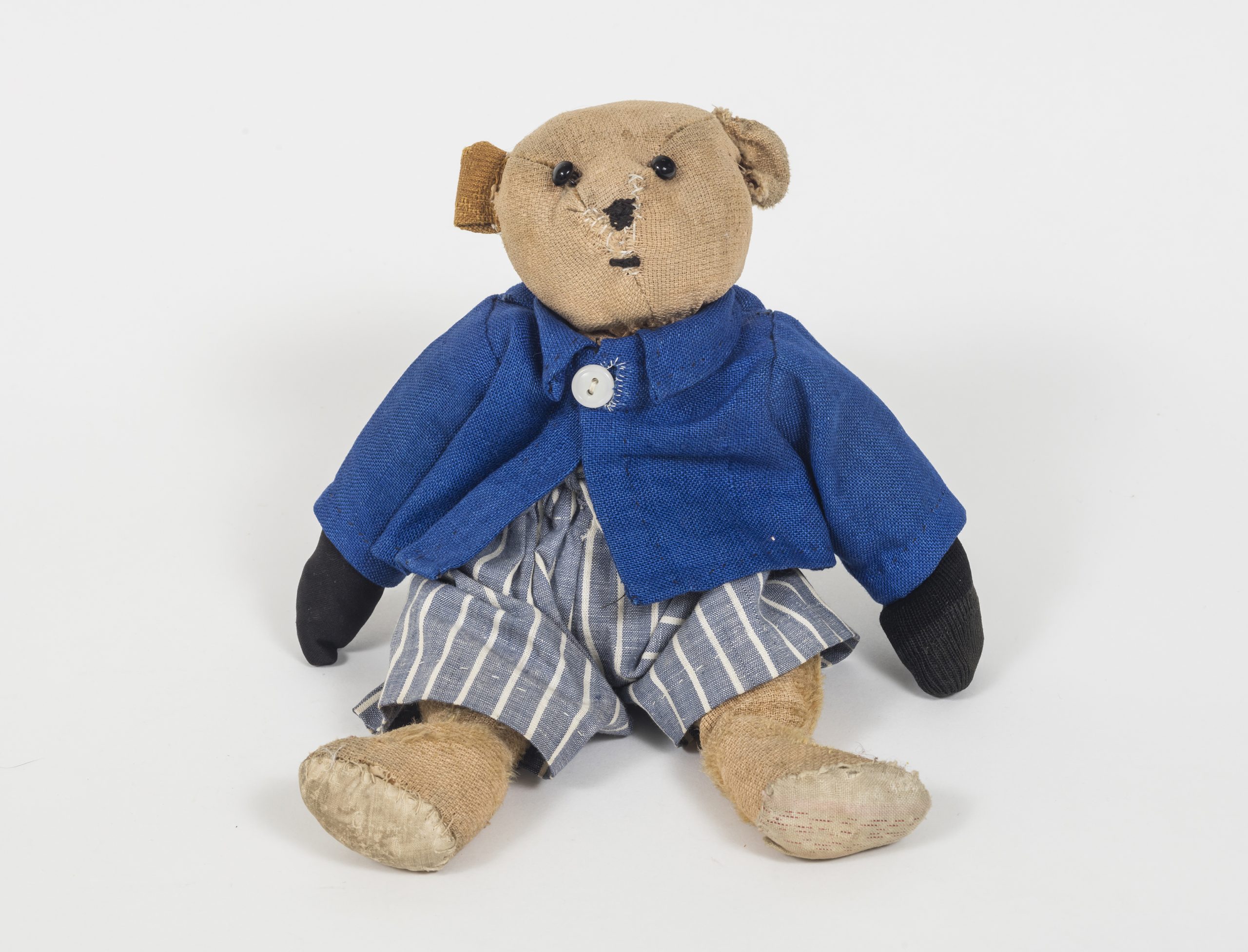 Teddy bear wearing a blue jacket, black mittens and blue and white striped pants