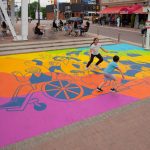 Children run across an outdoor square that has a colourful mural painted on the ground.