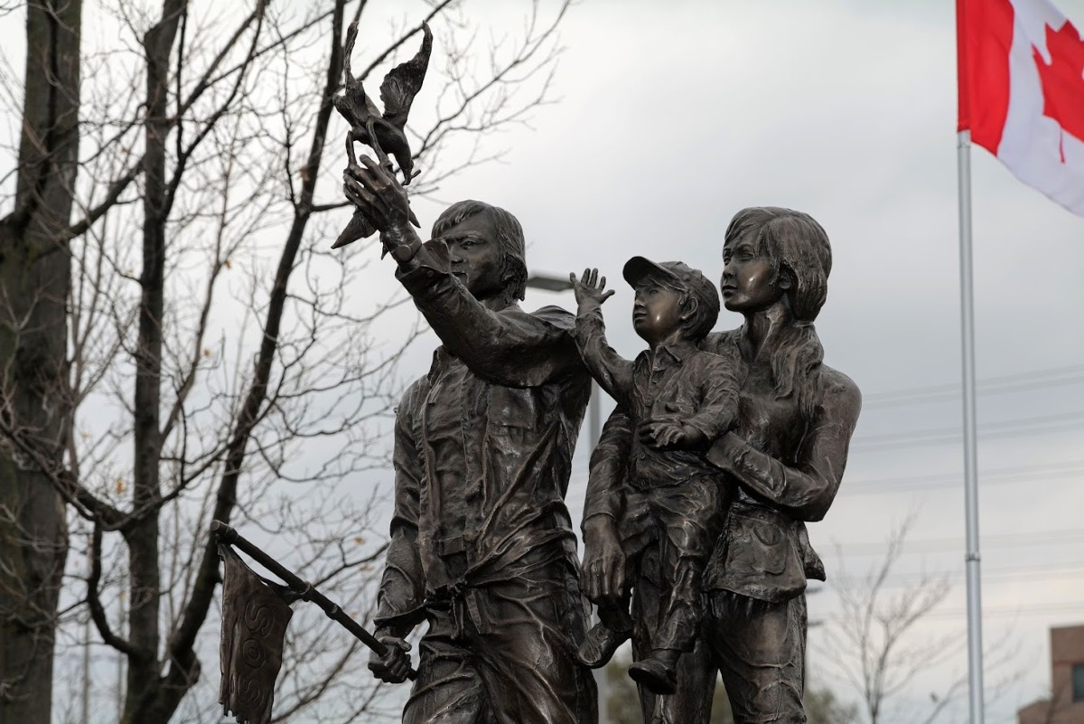 Bronze statue of a woman holding a child, and a man with his hand outstretched. A bird carrying a leaf sits on his hand. All three figures stand on a boat.