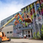 A construction crane lifts people up to paint a part of a mural at the top of a building.