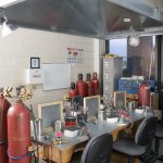 Rows of gas cylinders next to a flameworking table made of metal sheets and a wooden base. A large exhaust hood hangs overtop.