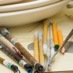 Paint brushes, pencils and other art tools on a wooden tabletop.