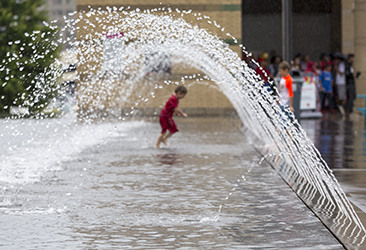 A young boy running through a wading pool with water spraying over his head.