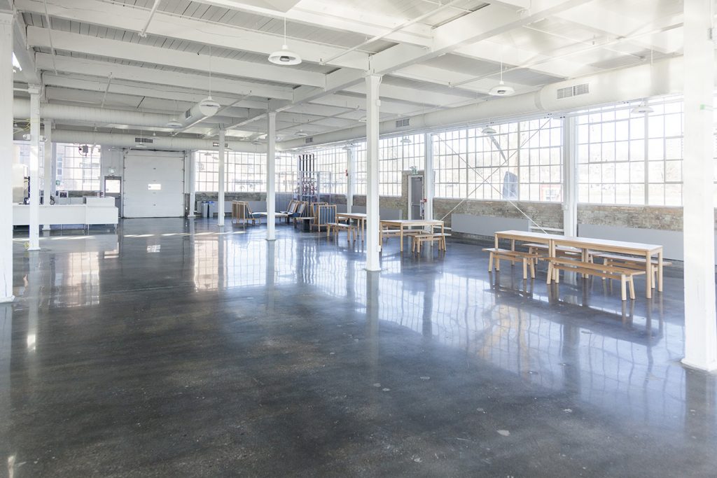 Brightly lit industrial space with rows of tables and chairs along one wall. CAPTION
