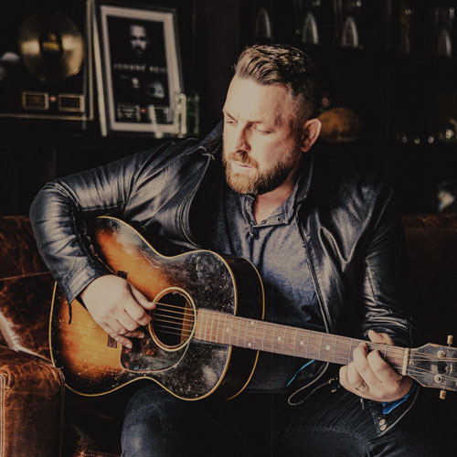 Singer Johnny Reid holding a guitar while sitting on a brown couch.