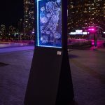 A neon art installation at night, featuring a luminous blue outline of a tiger and several flowers within a vertical frame, mounted on a slanted black pedestal. The backdrop reveals a cityscape with high-rise buildings, a tree's silhouette, and ambient city lights.