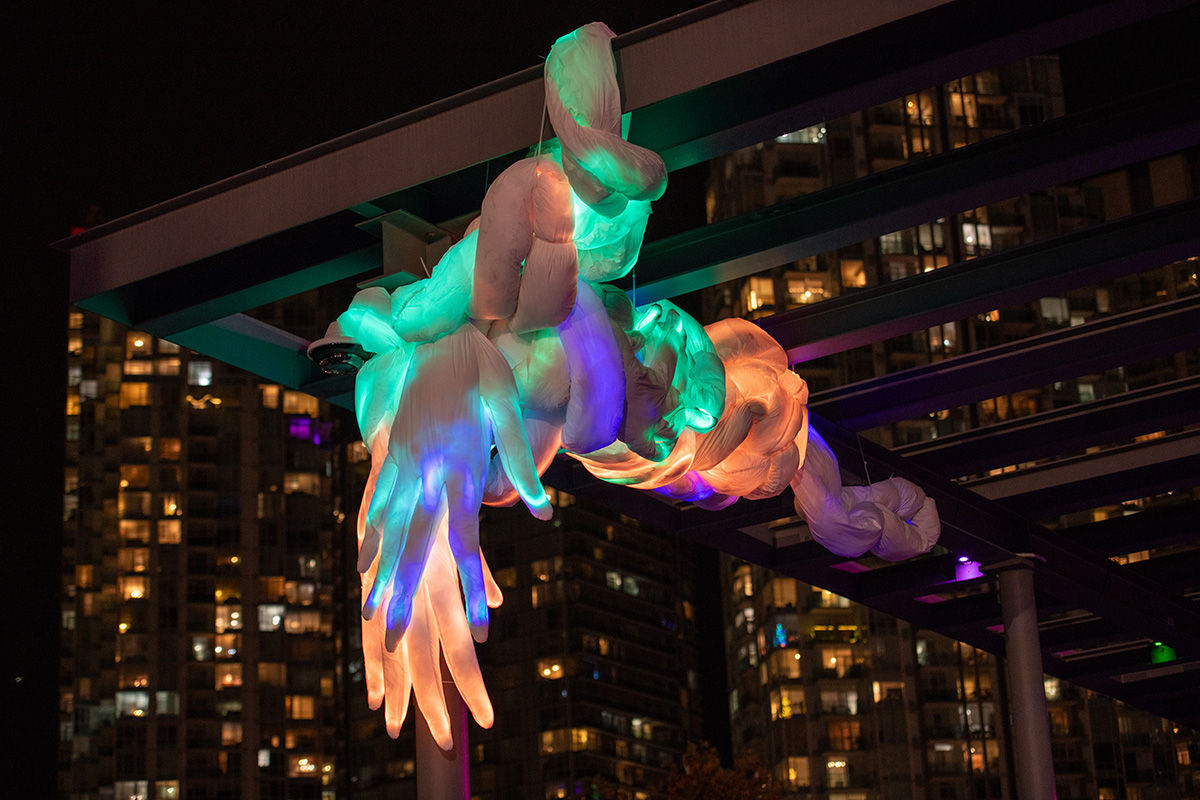 A sculptural light installation suspended from an overhead beam, featuring clustered, translucent shapes that glow in hues of blue, green, and pink, set against a night sky with the silhouette of high-rise apartment buildings in the background.