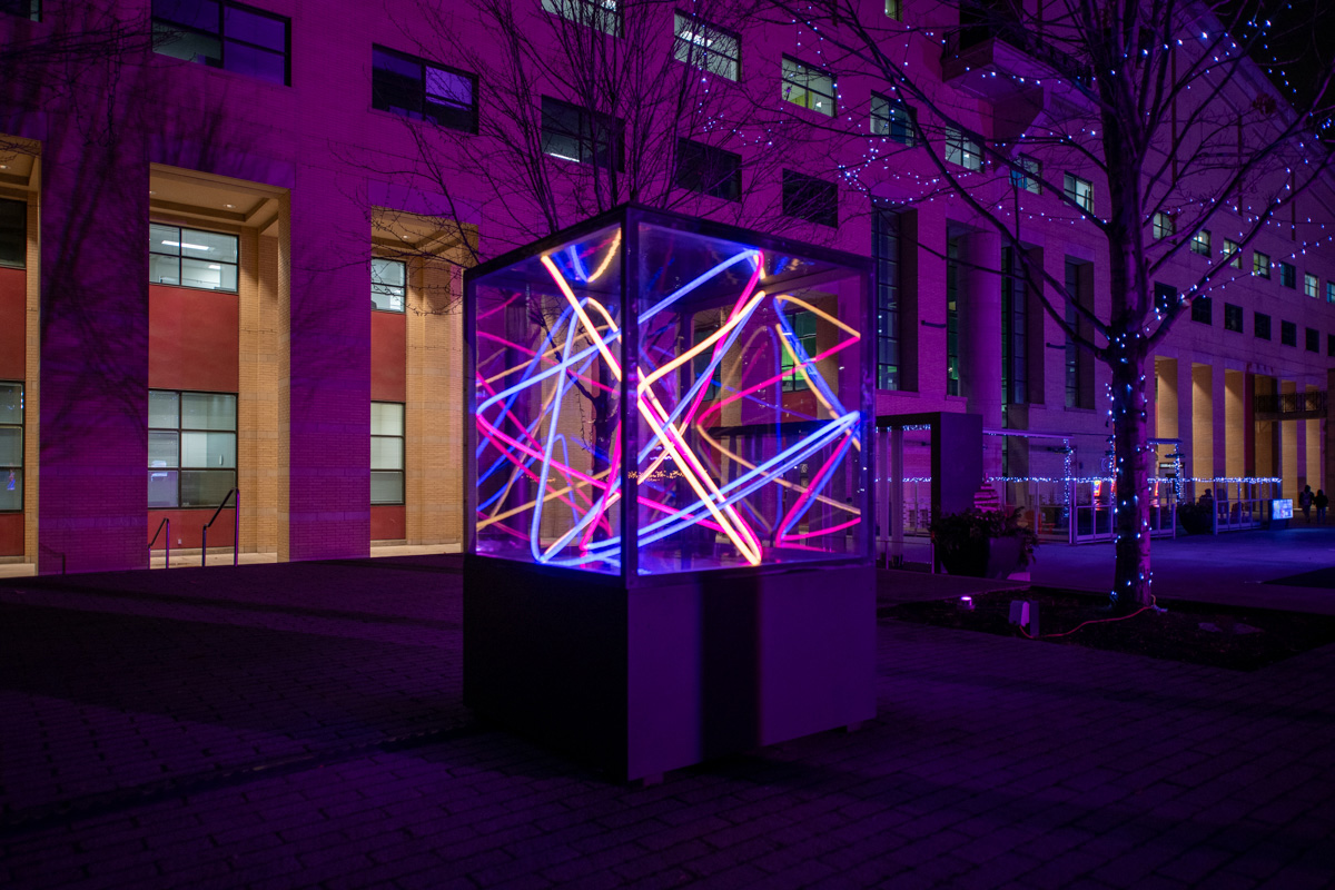 An outdoor art installation captured at dusk featuring a geometric structure with illuminated white lines forming a complex network within a transparent cube.