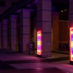 Neon light panels affixed to brick pillars along an outdoor walkway at night. The panels glow with vibrant geometric patterns in a sequence of colours, casting a colourful reflection on the pavement.