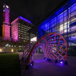 An illuminated architectural sculpture at night in an urban park, resembling a half-tunnel with a lattice structure. It is lit from below with blue and red lights, creating a striking contrast against the dark sky and the surrounding city buildings.