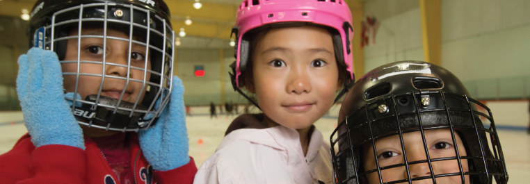 Three children wearing ice skating helmets inside a skating rink