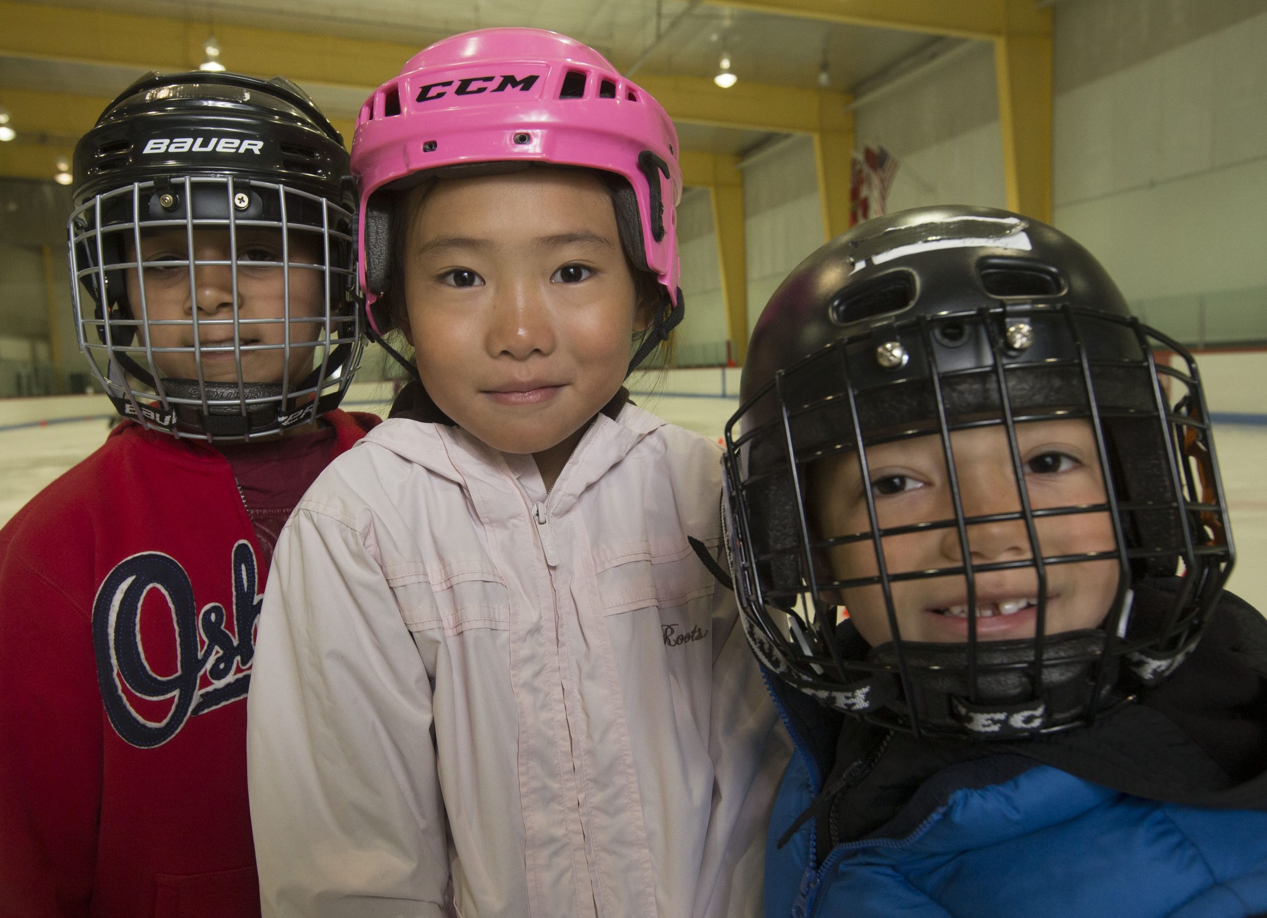 Photo: three children wearing hockey helmets inside an ice skating rink.