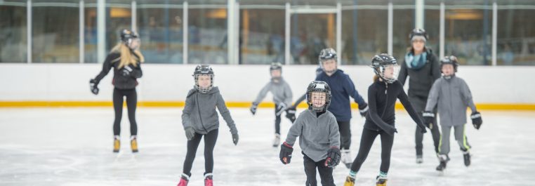 A group of kids are skating at an indoor rink. They are wearing helmets for safety.