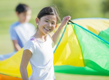 A girl is lifting a parachute and smiling at the camera.