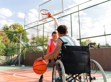 Teenage boy in wheelchair playing basketball with friend on sports court