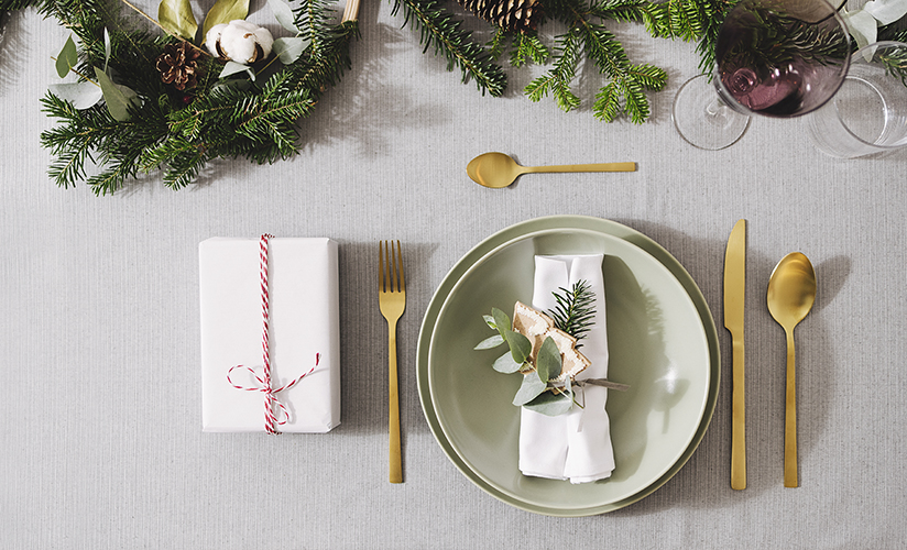 Flat lay of a holiday table setting with golden cutlery, green porcelain plate and present on a gray table.